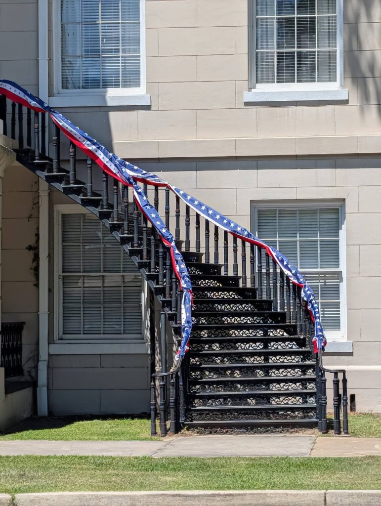 Black metal spiral staircase with a red, white, and blue patriotic banner, attached to a beige building with large windows.