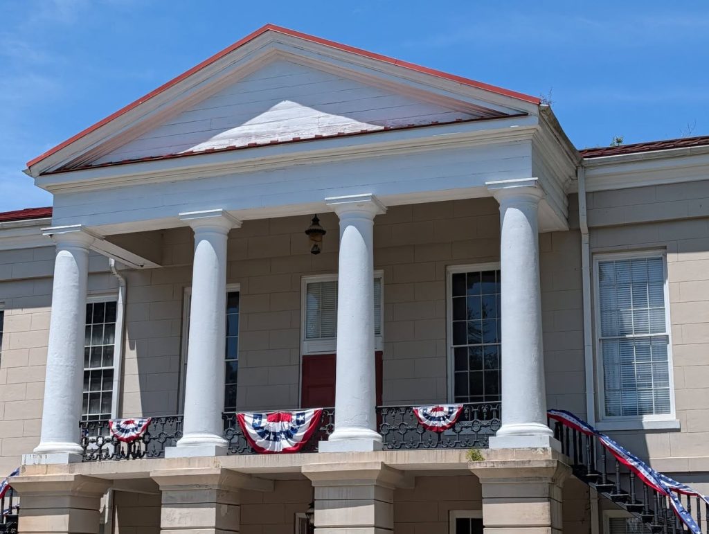 A building with four white columns, red door, and patriotic red, white, and blue banners on the balcony railings under a clear blue sky.