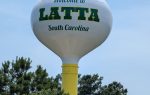 A white and yellow water tower reading "Welcome to Latta, South Carolina" rises above green trees against a blue sky.