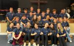 A group of young people and adults in matching blue shirts pose together indoors, smiling, with a wooden cross and musical instruments in the background.