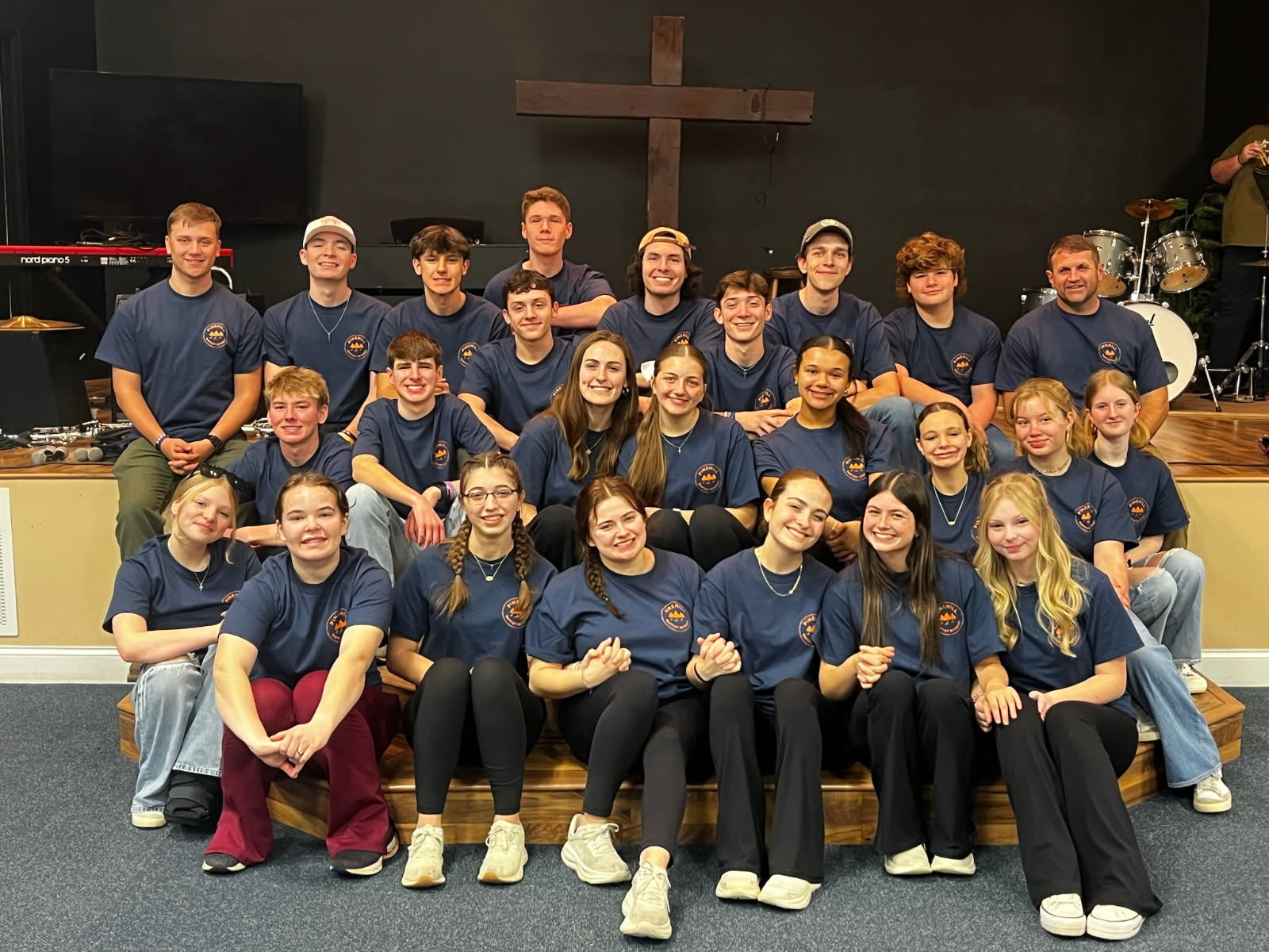 A group of young people and adults in matching blue shirts pose together indoors, smiling, with a wooden cross and musical instruments in the background.