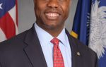 A man in a suit and tie smiles, standing in front of the American and South Carolina state flags.