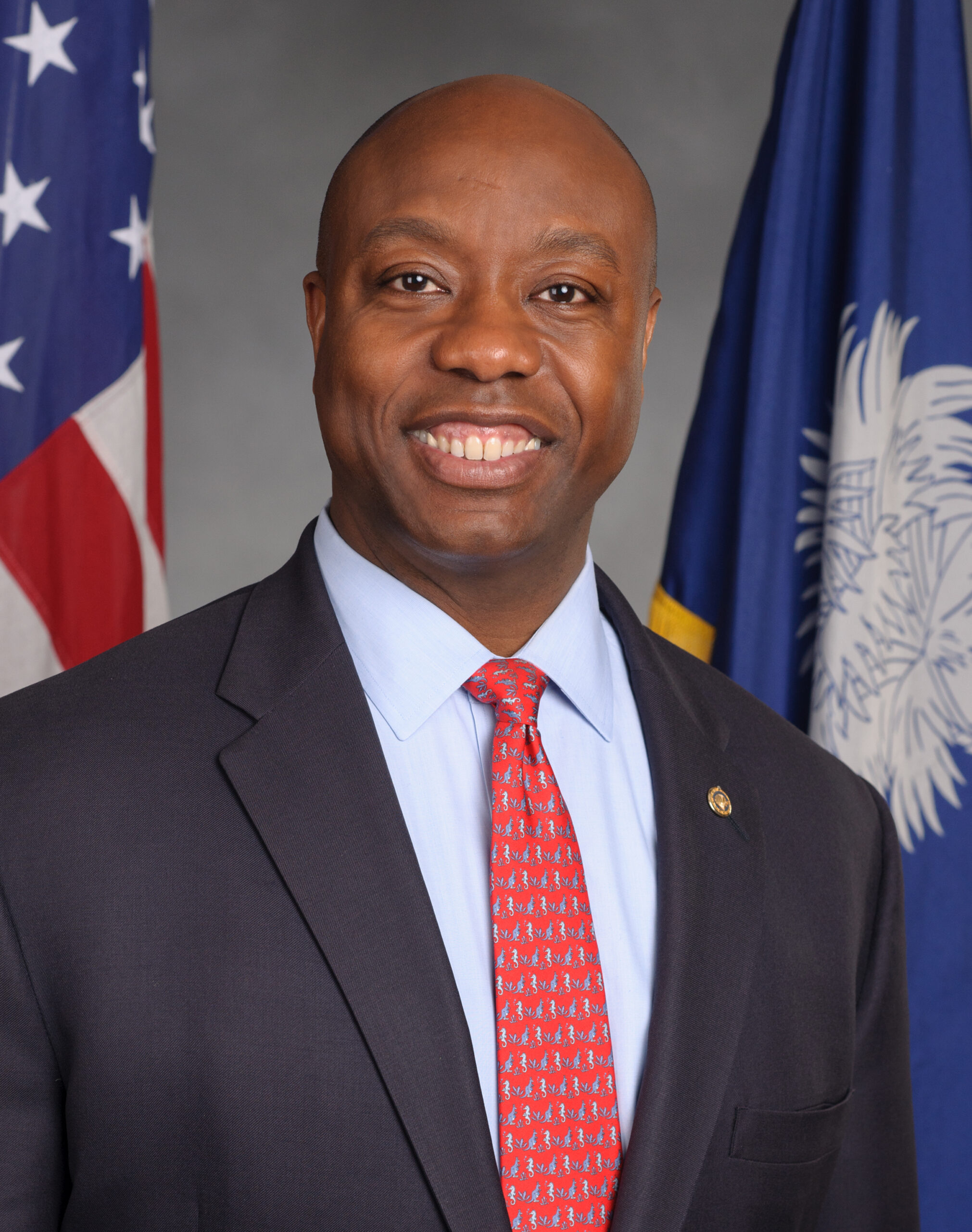A man in a suit and tie smiles, standing in front of the American and South Carolina state flags.