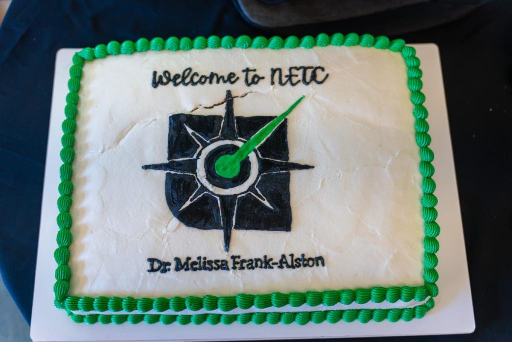 Rectangular cake with green icing border, compass design, and text reading "Welcome to NETC" and "Dr. Melissa Frank-Alston" on a white background.