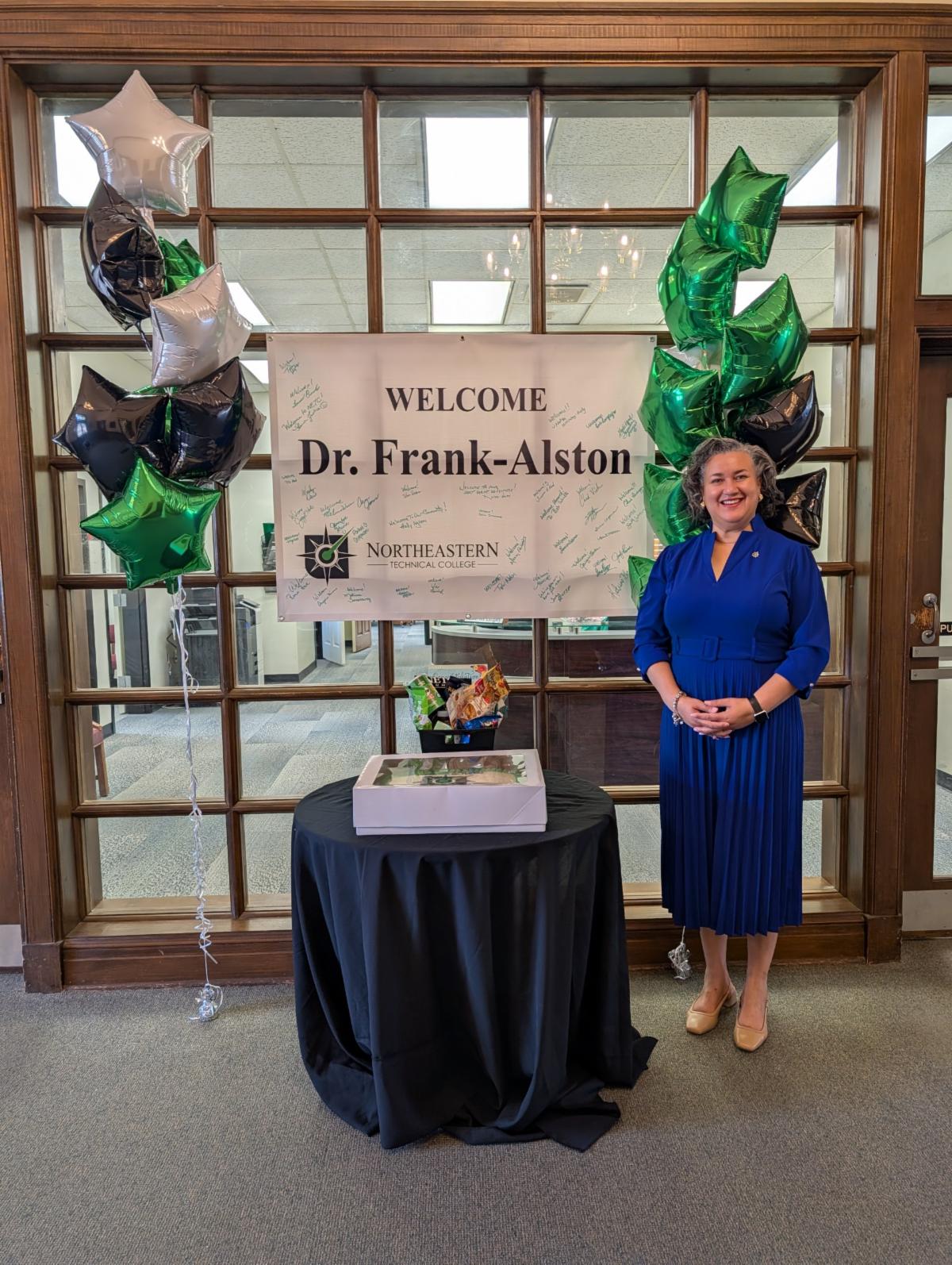 Woman standing beside a table and welcome sign for Dr. Frank-Alston at Northeastern Technical College, surrounded by green, black, and silver star-shaped balloons.