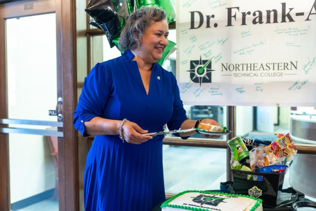 A woman in a blue dress smiles while serving cake at a celebration event with a Northeastern Technical College banner in the background.