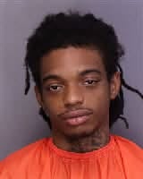 A young man with dreadlocks wearing an orange shirt poses against a plain background for a portrait-style photograph.