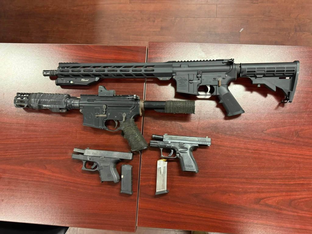 Four firearms, including two handguns and two rifles, are laid out on a wooden table.
