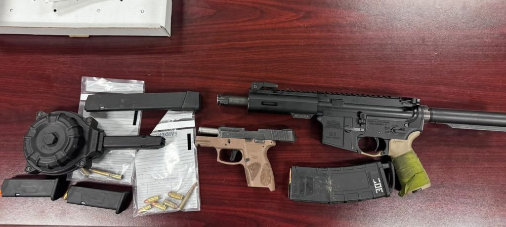 Two firearms, several magazines, loose bullets, and evidence bags displayed on a reddish wood table.