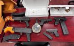 Various firearms, ammunition, and magazines displayed on a red wooden table, with cardboard boxes and empty ammo containers in the background.