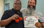 Two men smiling, one holding ripe tomatoes, the other displaying a "Tomato Growers Contest 2026 First Place" certificate in a casual indoor setting.