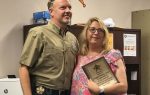 A man and woman stand together indoors, smiling, with the woman holding a plaque; office furniture and sheriff’s badge emblem are visible in the background.