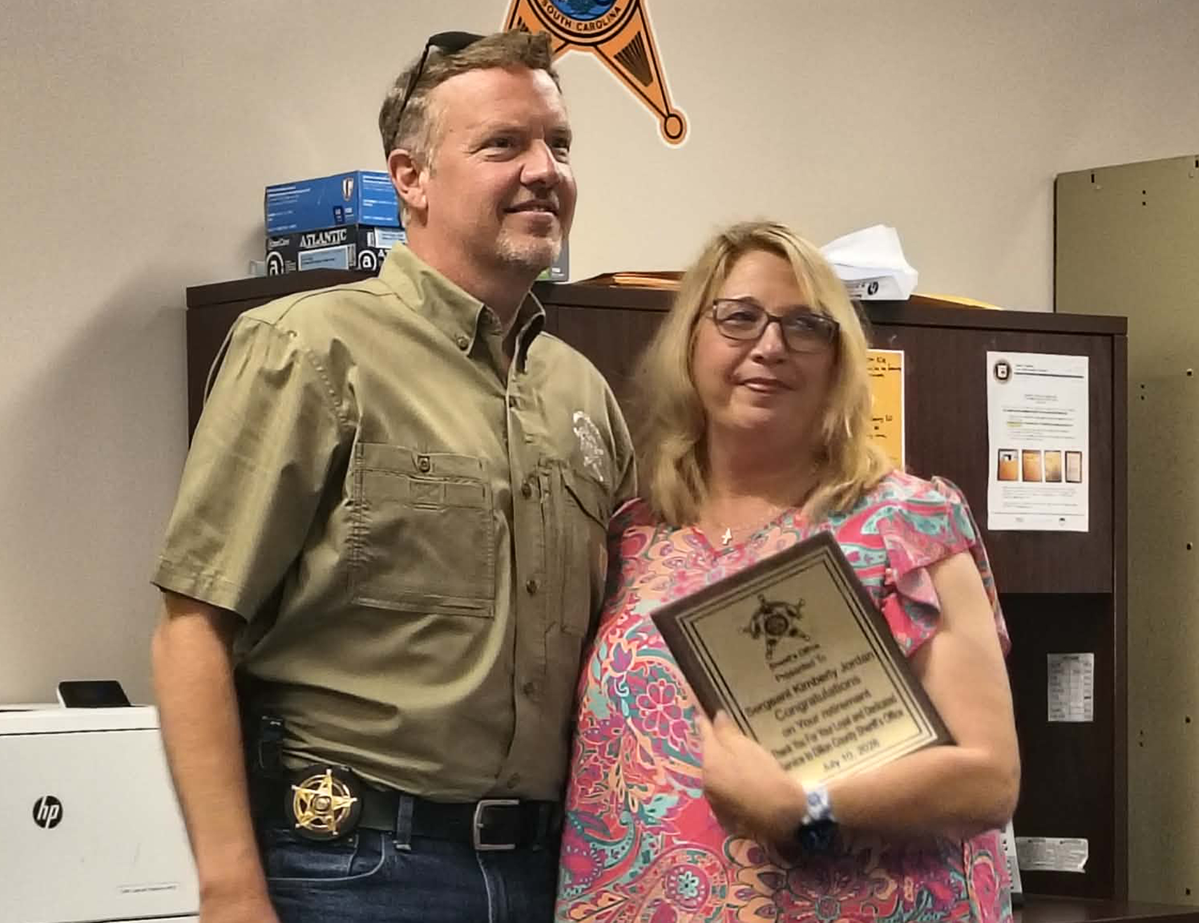 A man and woman stand together indoors, smiling, with the woman holding a plaque; office furniture and sheriff’s badge emblem are visible in the background.