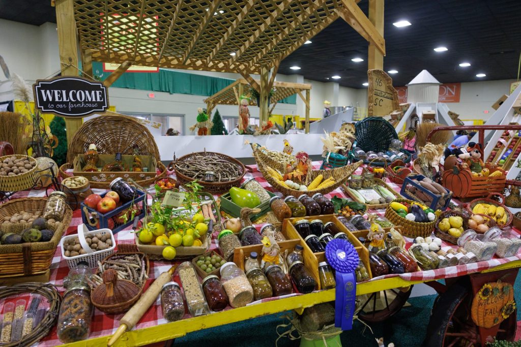 Farmers market display with fruits, vegetables, preserves, nuts, and homemade goods on a checkered table, under a wooden canopy, with a "Welcome to our farmhouse" sign.