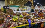 Farmers market display with fruits, vegetables, preserves, nuts, and homemade goods on a checkered table, under a wooden canopy, with a "Welcome to our farmhouse" sign.