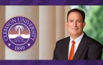 A man in a suit and orange tie poses beside the Clemson University seal, which displays "Clemson University, South Carolina, 1889."