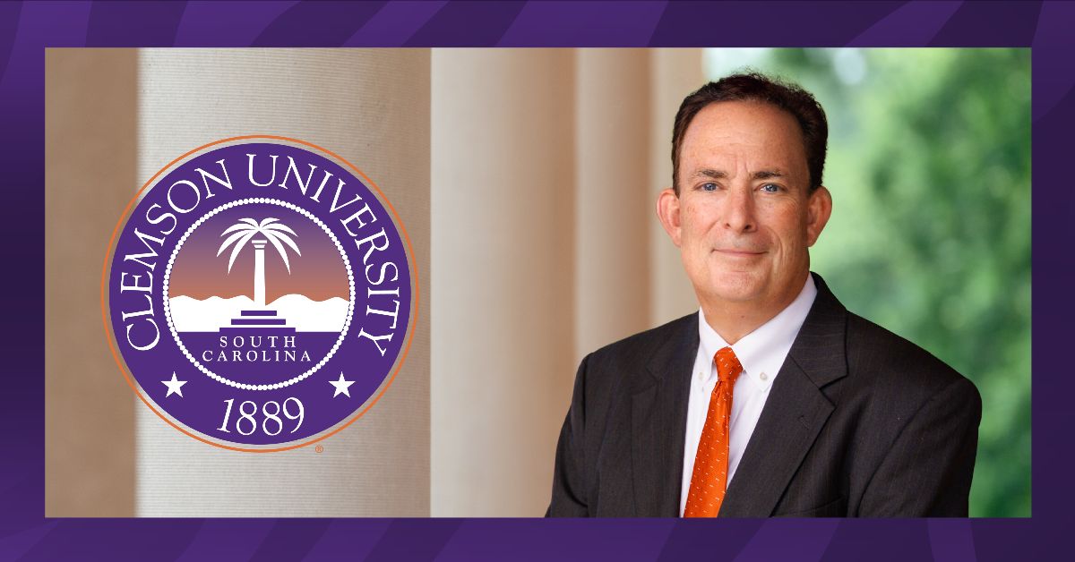 A man in a suit and orange tie poses beside the Clemson University seal, which displays "Clemson University, South Carolina, 1889."